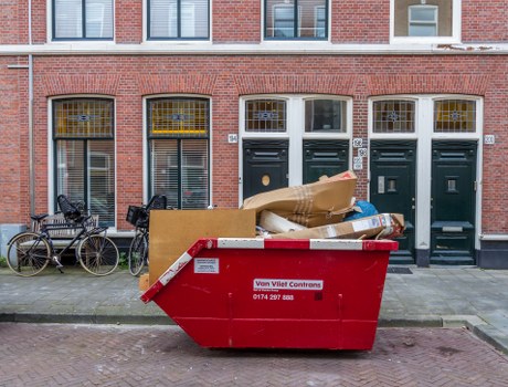 A large red skip bin placed on a paved urban street in front of a row of three storey brick terraced houses with dark green doors and white framed windows. The skip is filled with various waste materials, including large cardboard boxes, plastic bags, and loose rubbish, some partially overhanging the edge. To the left of the skip, there are two bicycles parked against the street pavement near the building, one with a black frame and the other with a silver frame. The background features a brick façade with a clean, orderly appearance, and the street appears quiet with no pedestrians visible. The scene suggests a rubbish removal or clearance service in Kilburn, with the skip serving as a temporary container for waste collection, typical of local garden or household clearance activities carried out by Garden Clearance Kilburn.