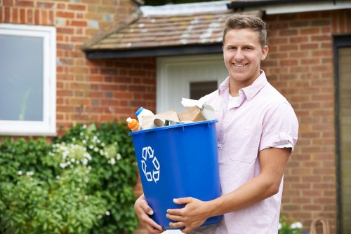 A young man with short light brown hair and a friendly smile stands outdoors in front of a brick house with a white door and a window, holding a large blue recycling bin filled with various rubbish items such as crumpled paper, plastic bottles, and cardboard boxes. He is wearing a light pink short-sleeved shirt, and the bin features a white recycling symbol on its side. Behind him, there is a lush green hedge with small white flowers, and the setting appears to be a residential driveway or front garden in Kilburn, suggesting a professional rubbish or garden clearance service. The overall scene is well-lit with natural daylight, emphasizing the tidy and orderly environment, consistent with the offerings of Garden Clearance Kilburn for local rubbish removal.