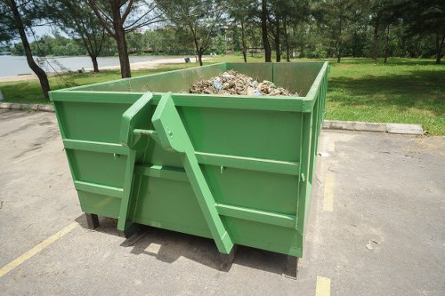 A large green metal skip filled with a mixture of garden debris, including twigs, leaves, and soil, positioned on a paved area adjacent to a grassy parkland with trees in the background. The skip features reinforced sides with horizontal ribs and is situated in an outdoor setting typical of garden clearance work near Kilburn, with the surrounding environment consisting of a paved parking space and a lawn with mature trees. The scene is lit with natural daylight, emphasizing the textures of the metal and organic waste, and is representative of rubbish removal services provided by Garden Clearance Kilburn for garden waste disposal in the local area.