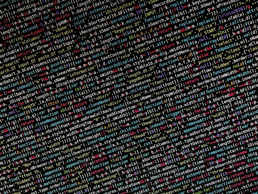 A close-up photograph of a laptop placed on a wooden surface, with visible lines of computer code displayed on the screen. The code includes syntax highlighting in green, blue, and white on a dark bac