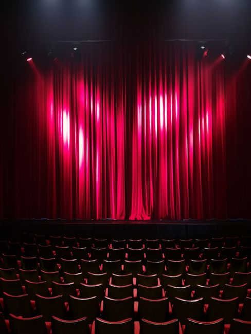 An empty theatre auditorium featuring a closed stage with red velvet curtains hanging from ceiling to floor. The curtains are illuminated by stage lighting, creating a glossy texture and a vibrant pin