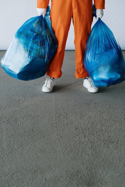 A person wearing orange work overalls and white sneakers stands on a grey floor, holding two large blue plastic rubbish bags filled with waste materials. The bags appear to be made of semi-transparent